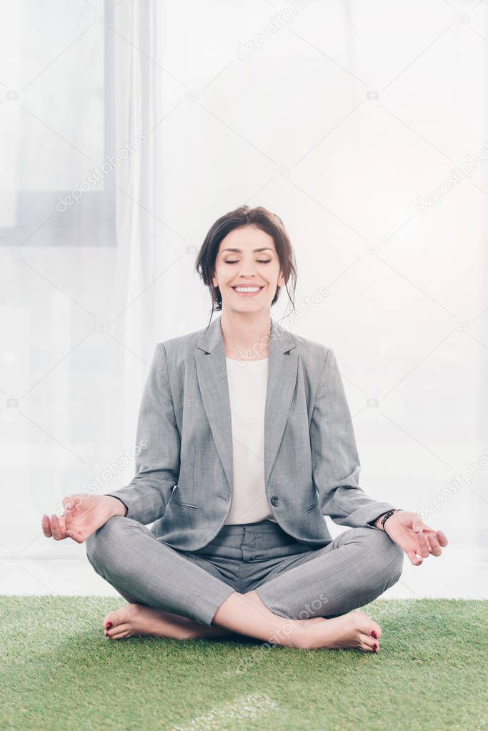beautiful smiling businesswoman in suit meditating while sitting on grass mat in Lotus Pose 