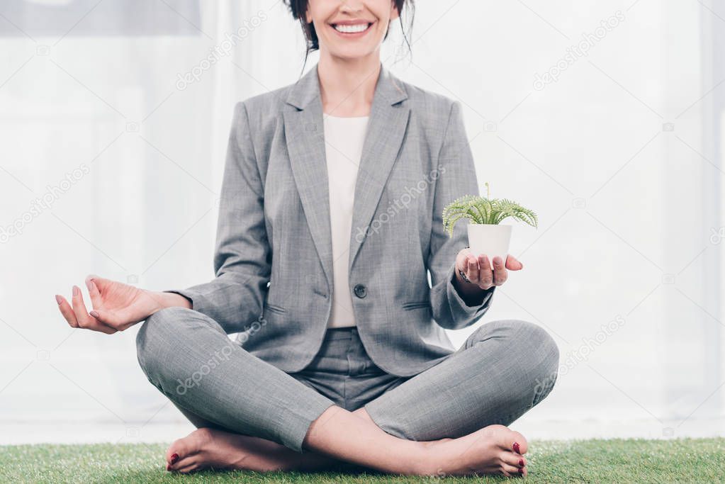 cropped view of smiling businesswoman in suit meditating on grass mat in Lotus Pose with flowerpot 