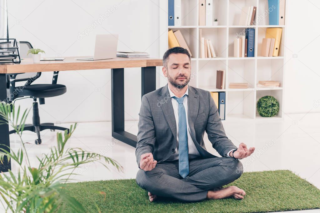 businessman sitting on grass mat in Lotus Pose and meditating in office