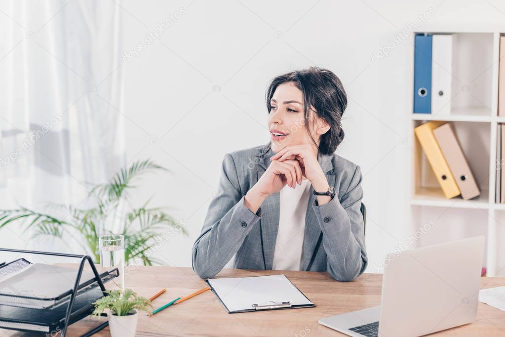 beautiful businesswoman in suit sitting at table with clipboard and laptop in office 