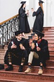 selective focus of cheerful students in graduation gowns holding diplomas and paper cups while sitting on stairs 