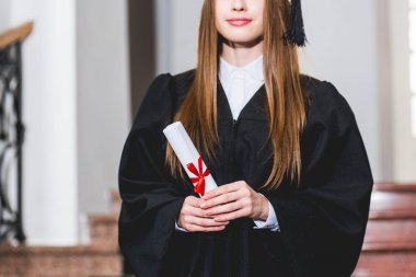 üniversitede diploma tutan ve gülümseyen neşeli genç kadının kırpılmış görünümü 