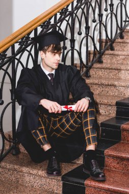 mezuniyet elbisesi içinde yakışıklı öğrenci merdivenlerde oturan ve diploma tutan 