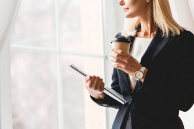 pencerenin yanında duran ve kağıt bardak ve klasör tutan sarışın öğretmen kırpılmış görünümü 