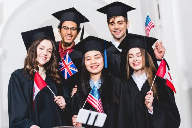 farklı ülkelerin bayraklarını tutan ve akıllı telefonda selfie çekmek mezuniyet önlüklerinde neşeli öğrencilerin seçici odak 