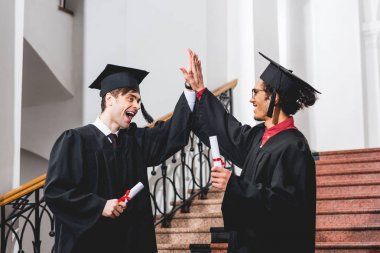 mezuniyet kapaklarında neşeli öğrenciler yüksek beş veren diplomaları tutarken 
