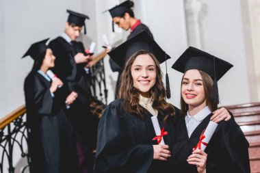 öğrencilerin yanında diploma tutarken mezuniyet kapakları gülümseyen güzel kızların seçici odak