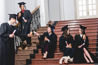 kağıt bardak ve diplomaile merdivenlerde oturan mezuniyet önlük öğrencilerin neşeli grup 