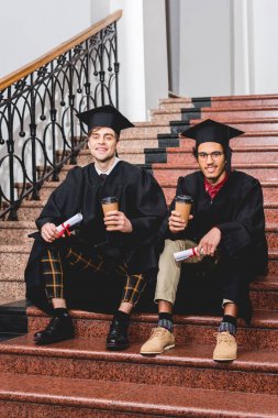 mezuniyet önlük neşeli öğrenciler merdivenlerde otururken diploma ve kağıt bardak tutan 