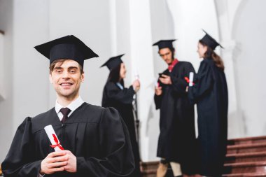 mezuniyet elbisesi içinde mutlu adam seçici odak kamera bakarak ve öğrencilerin yanında diploma tutarak 