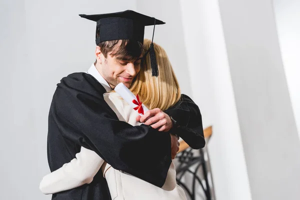 Cheerful Son Graduation Cap Holding Diploma While Hugging Blonde Mother — Stock Photo, Image