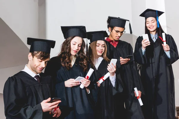 Cheerful Students Graduation Caps Using Smartphones While Holding Diplomas — Stock Photo, Image