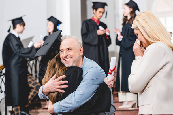 back view of girl in graduation cap hugging with father near crying mother