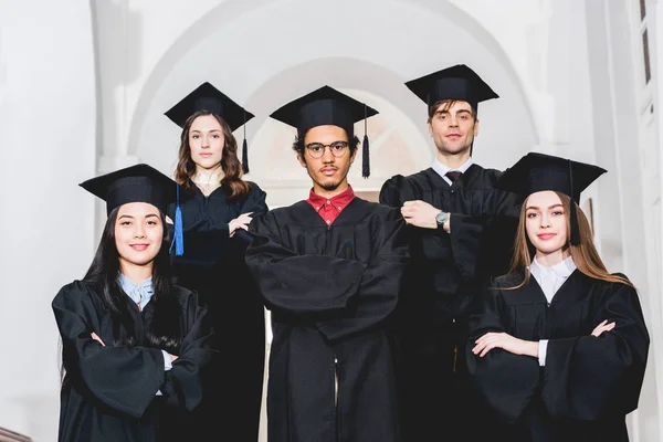 欢快的学生在毕业礼服站在交叉的手臂在大学 — 图库照片