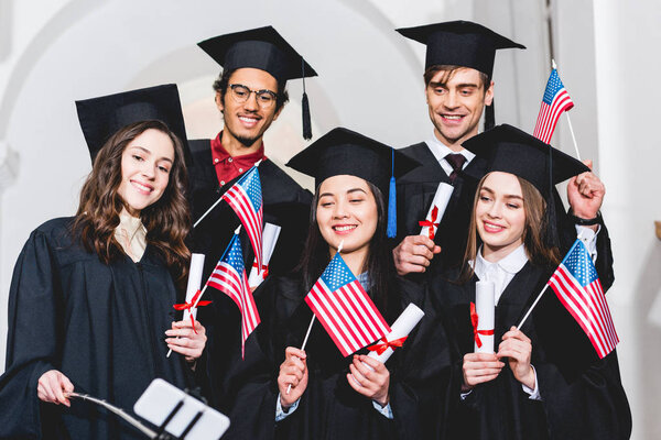 selective focus of happy students in graduation gowns holding flags of america and taking selfie on smartphone 