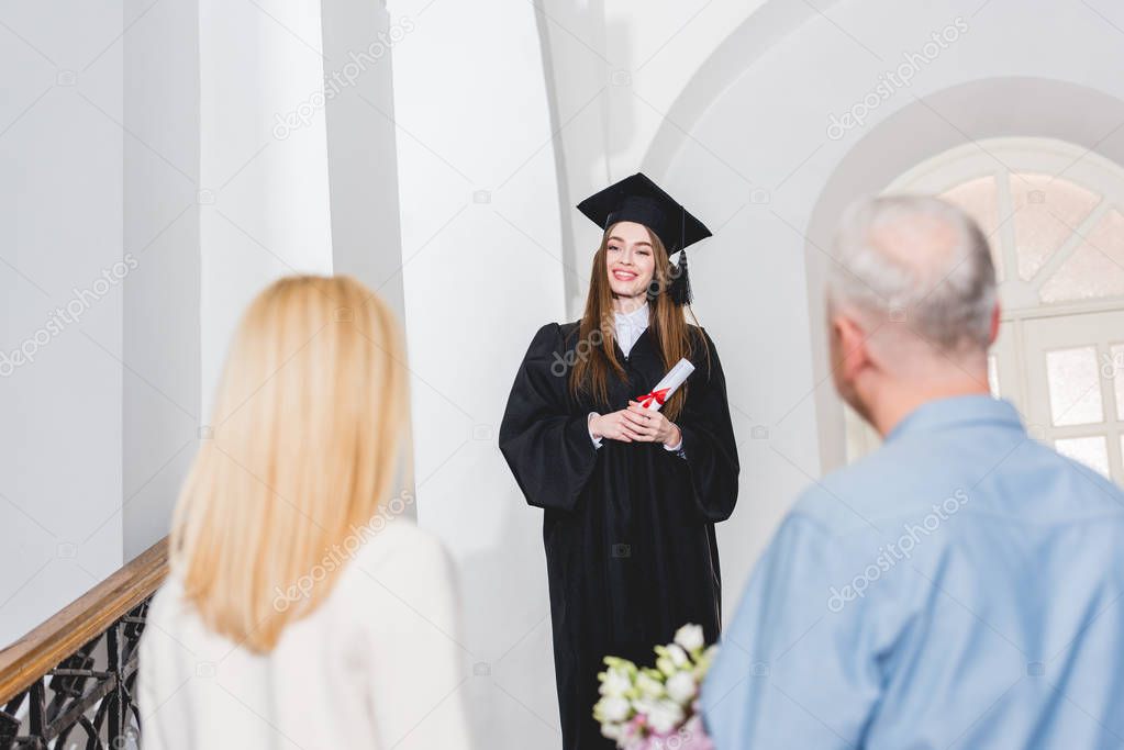 selective focus of cheerful girl in graduation cap looking at parents and holding diploma 