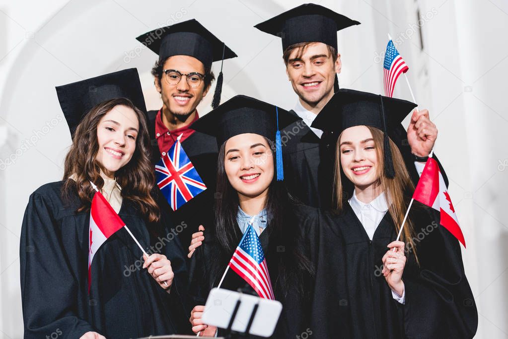 selective focus of cheerful students in graduation gowns holding flags of different countries and taking selfie on smartphone 