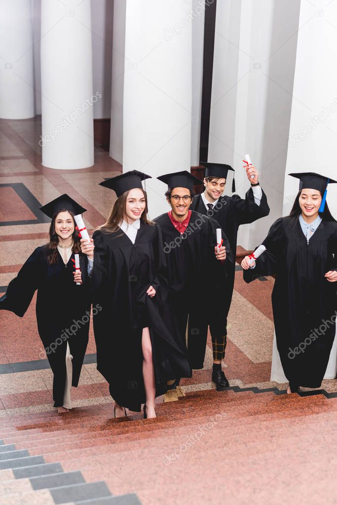 cheerful students in graduation gowns holding diplomas and walking on stairs 