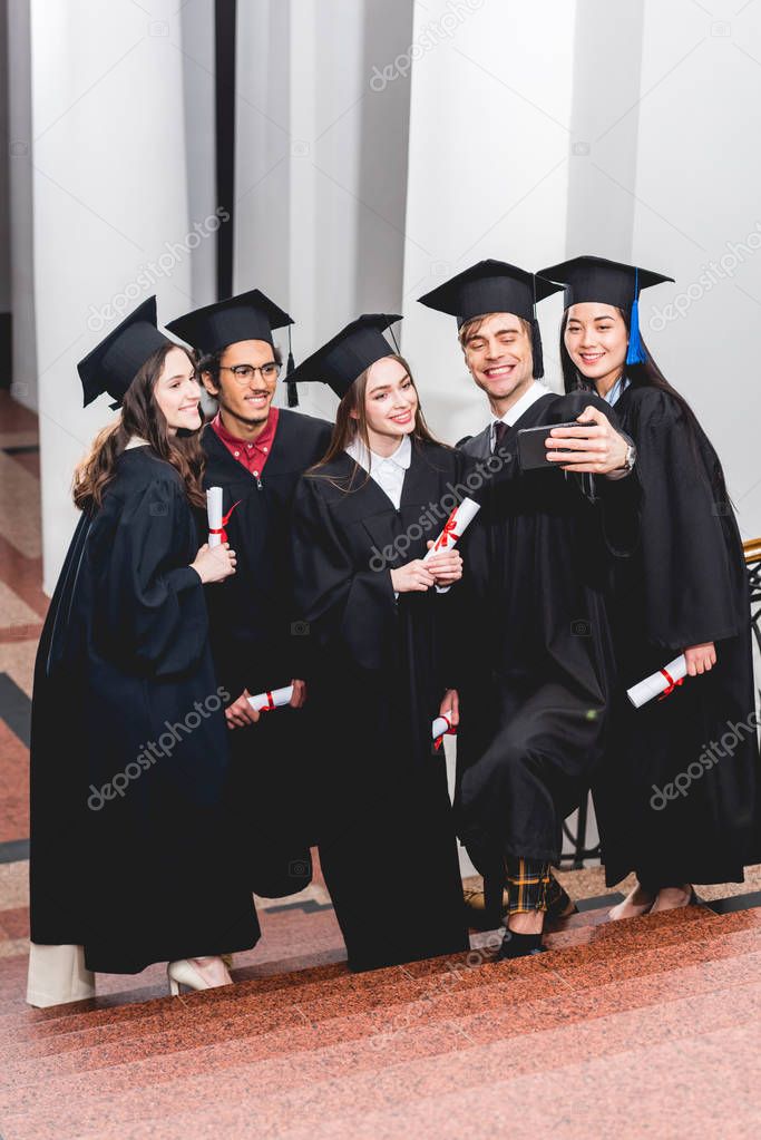 happy students in graduation gowns taking selfie and smiling while holding diplomas 