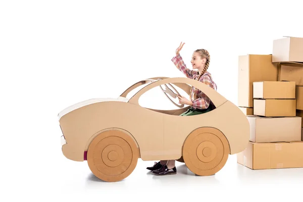 happy kid sitting and waving in cardboard car near packages on white