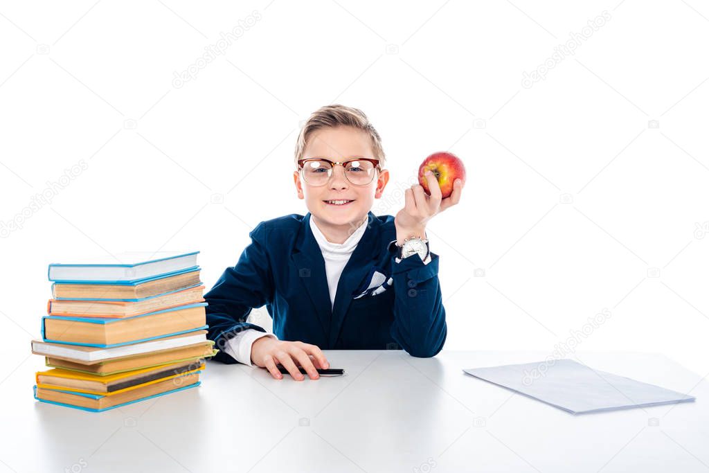 happy schoolboy in glasses sitting at desk with books and holding apple isolated on white