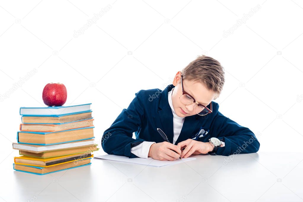 schoolboy in glasses sitting at desk with books and writing isolated on white