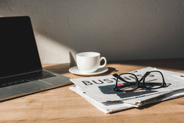 selective focus of glasses on business newspaper near laptop and cup with saucer on wooden table 