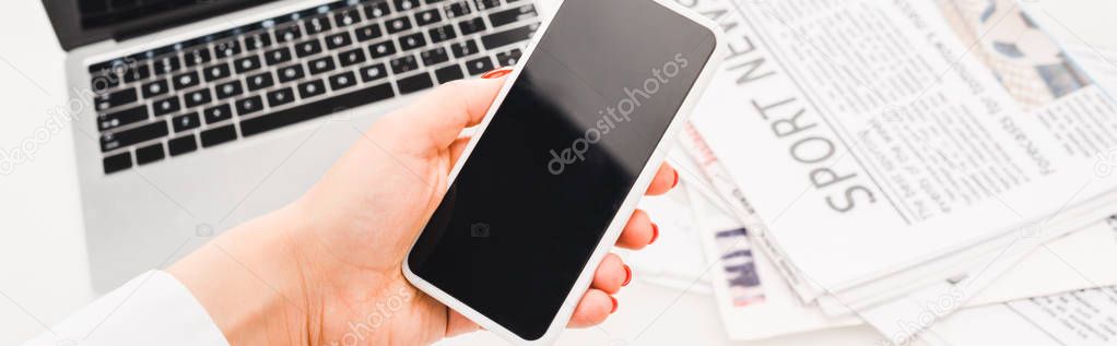 panoramic shot of woman holding smartphone with blank screen near laptop and  newspapers