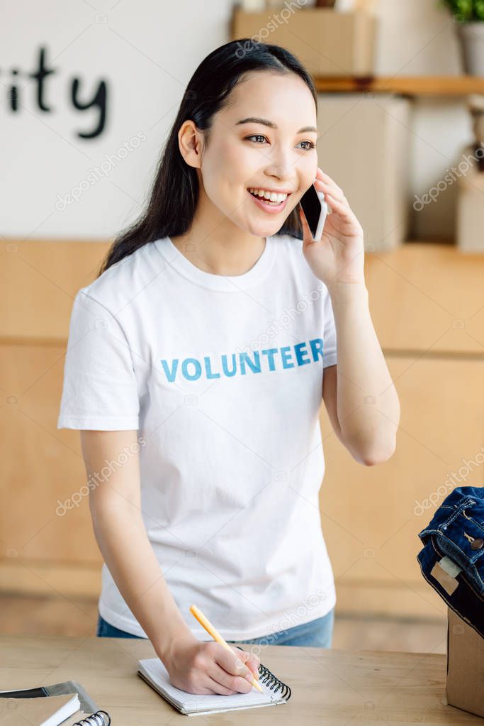 attractive asian volunteer talking on smartphone and writing in notebook