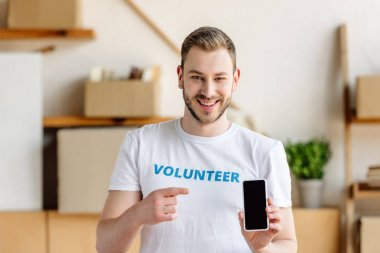 smiling young volunteer pointing with finger at smartphone with blank screen clipart