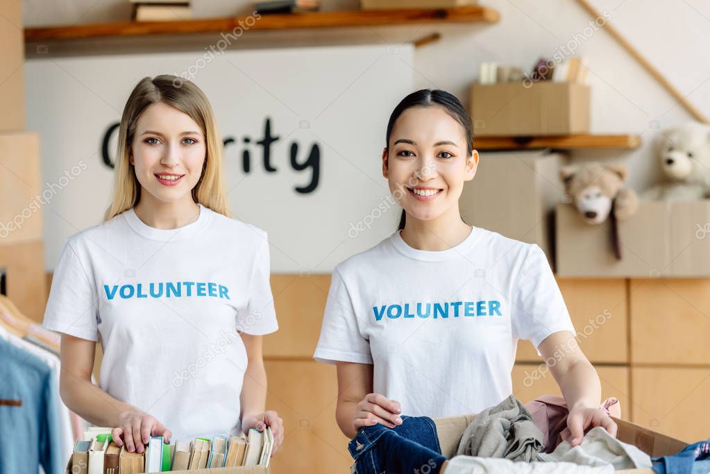 two pretty multicultural volunteers standing near carton boxes with books and clothes and looking at camera