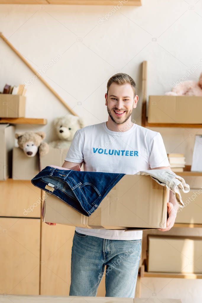 handsome, cheerful volunteer holding cardboard box with clothes
