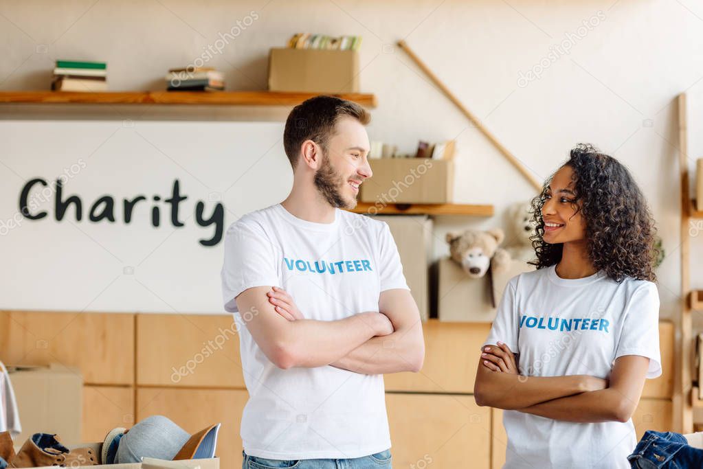 two cheerful multicultural volunteers standing with crossed arms, smiling and looking at each other