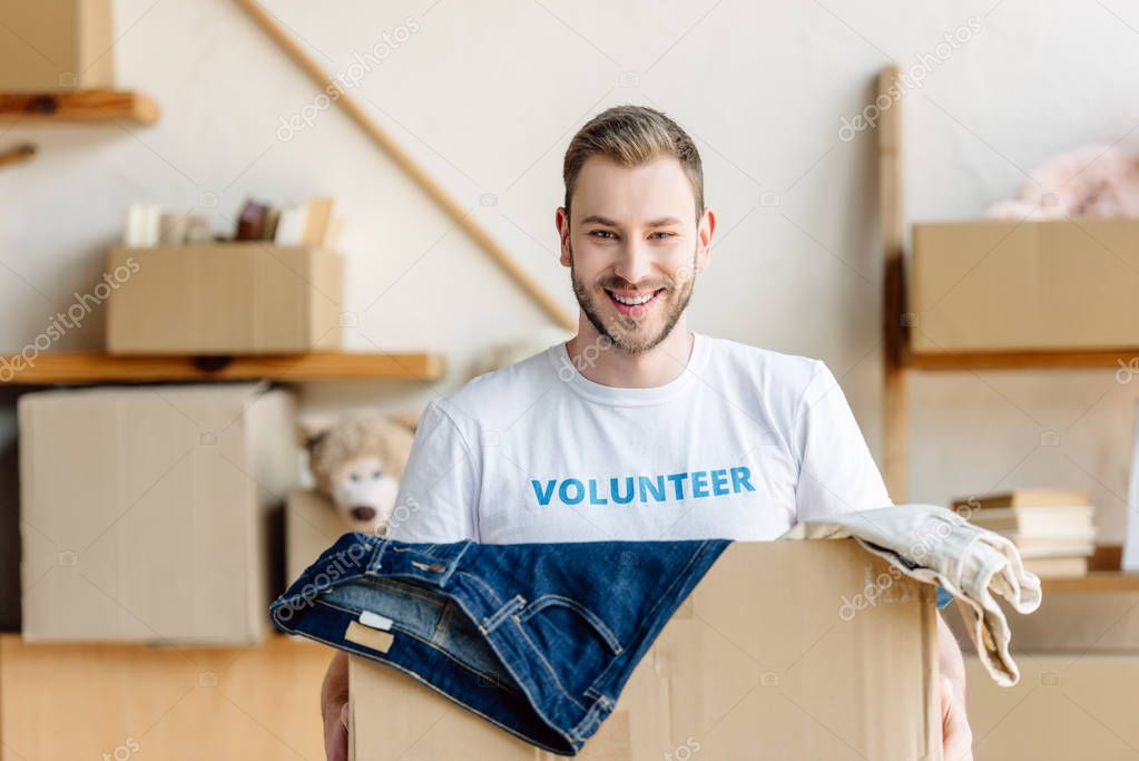 handsome young volunteer holding carton box with clothes, smiling and looking at camera