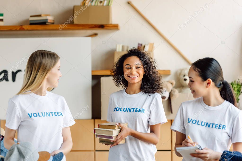 pretty, multicultural girls in white t-shirts with volunteer inscriptions smiling and looking at each other 