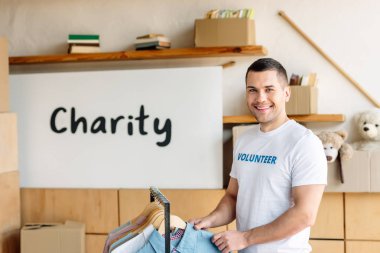 good-looking, cheerful volunteer standing near rack with shirts and looking at camera clipart