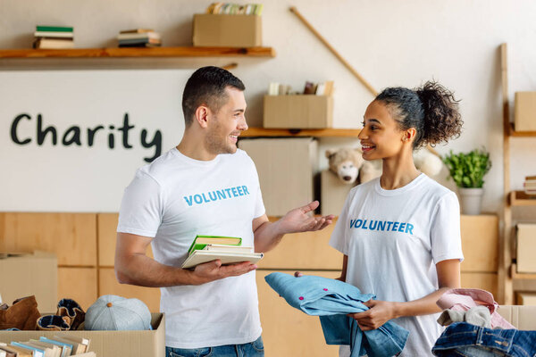 Два весёлых мультикультурных волонтера разговаривают, держа книги и одежду
