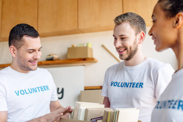 трое молодых мультикультурных волонтеров улыбаются и разговаривают, держа картонную коробку с книгами
