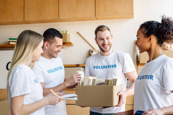 четыре молодых мультикультурных волонтера разговаривают, держа картонную коробку с книгами
