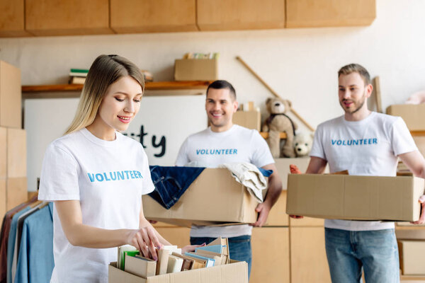 селективное внимание красивой девушки распаковывает коробку с книгами и красивых волонтеров держащих коробки с одеждой
