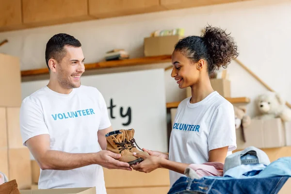 Two young, smiling multicultural volunteers holding childrens shoes in charity center — Stock Image Two Young Smiling Multicultural Volunteers Holding Childrens Shoes Charity Center Stock Photo