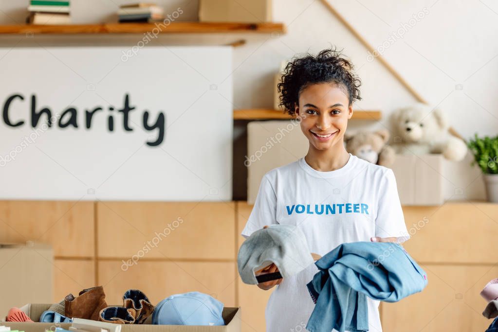 pretty african american volunteer holding clothes, smiling and looking at camera