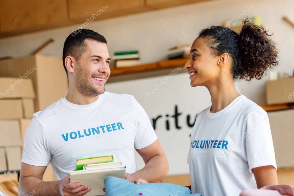 two cheerful multicultural volunteers talking while holding books and clothes