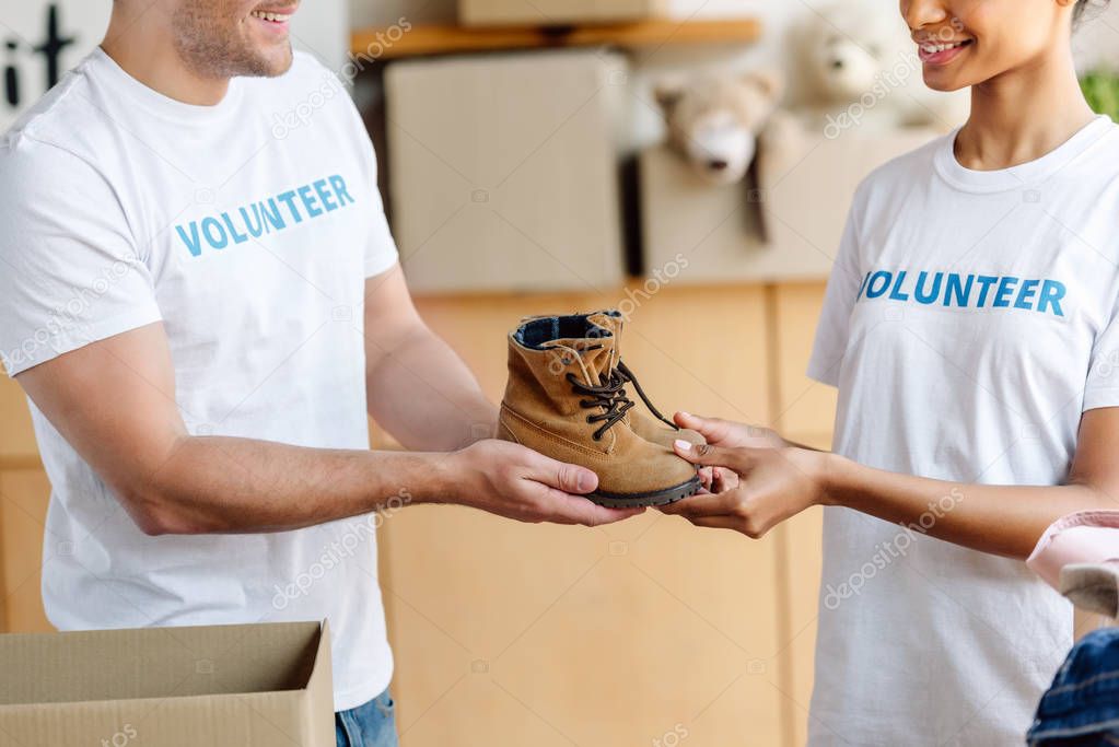 cropped view of two multicultural volunteers holding childrens shoes in charity center