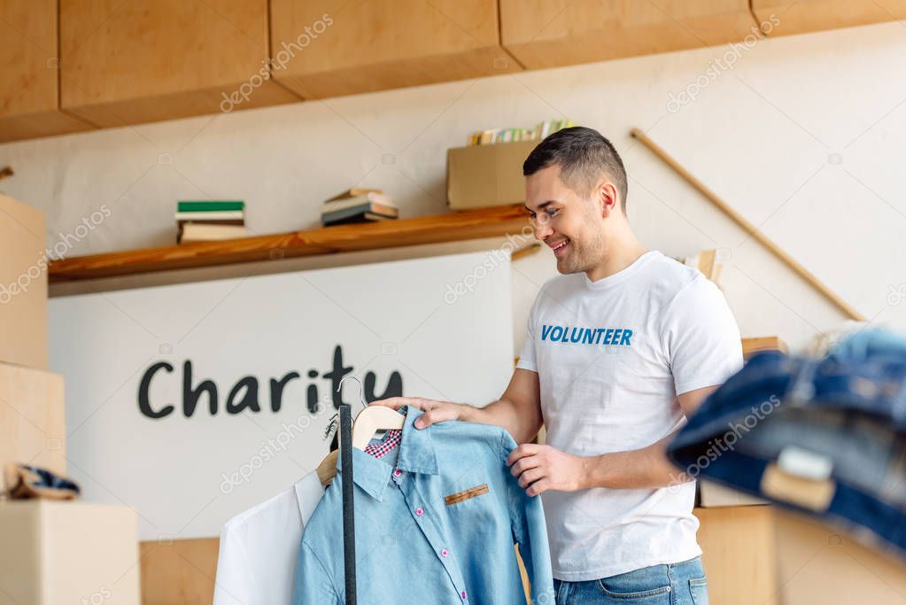 selective focus of handsome, smiling volunteer standing near rack with shirts