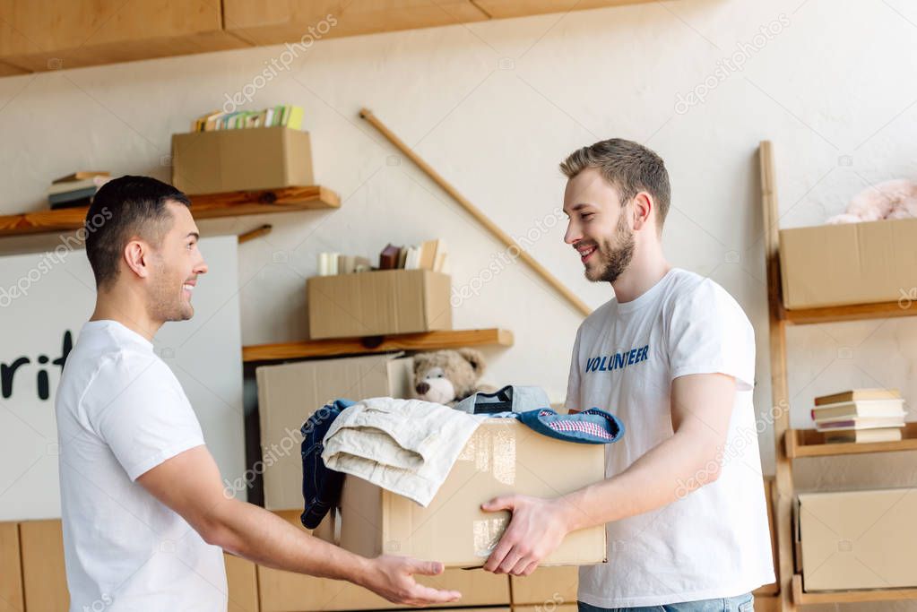 two good-looking, smiling volunteers holding cardboard box with clothes in charity center