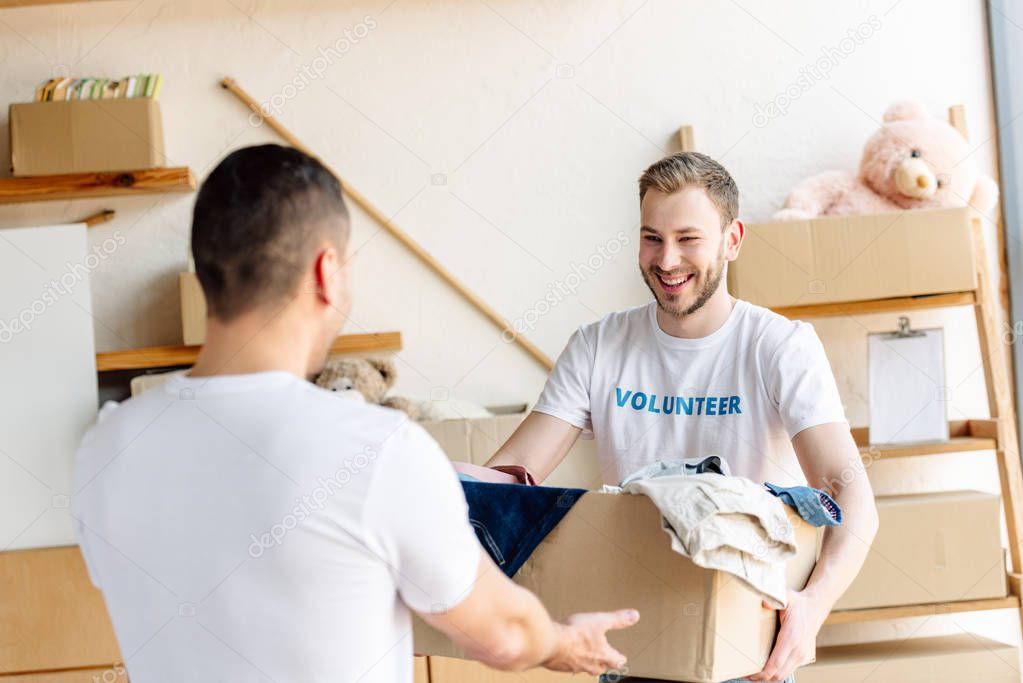two young volunteers holding cardboard box with clothes in charity center
