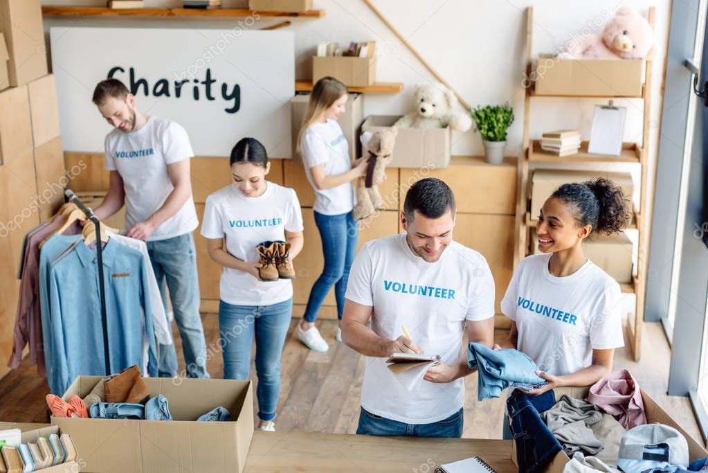 young, cheerful multicultural volunteers in white t-shirts with volunteer inscriptions working in charity center