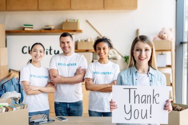 selective focus of pretty girl holding placard with thank you text while standing near multicultural volunteers in charity center clipart