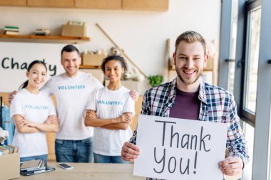 selective focus of handsome man holding placard with thank you text while standing near multicultural volunteers in charity center clipart